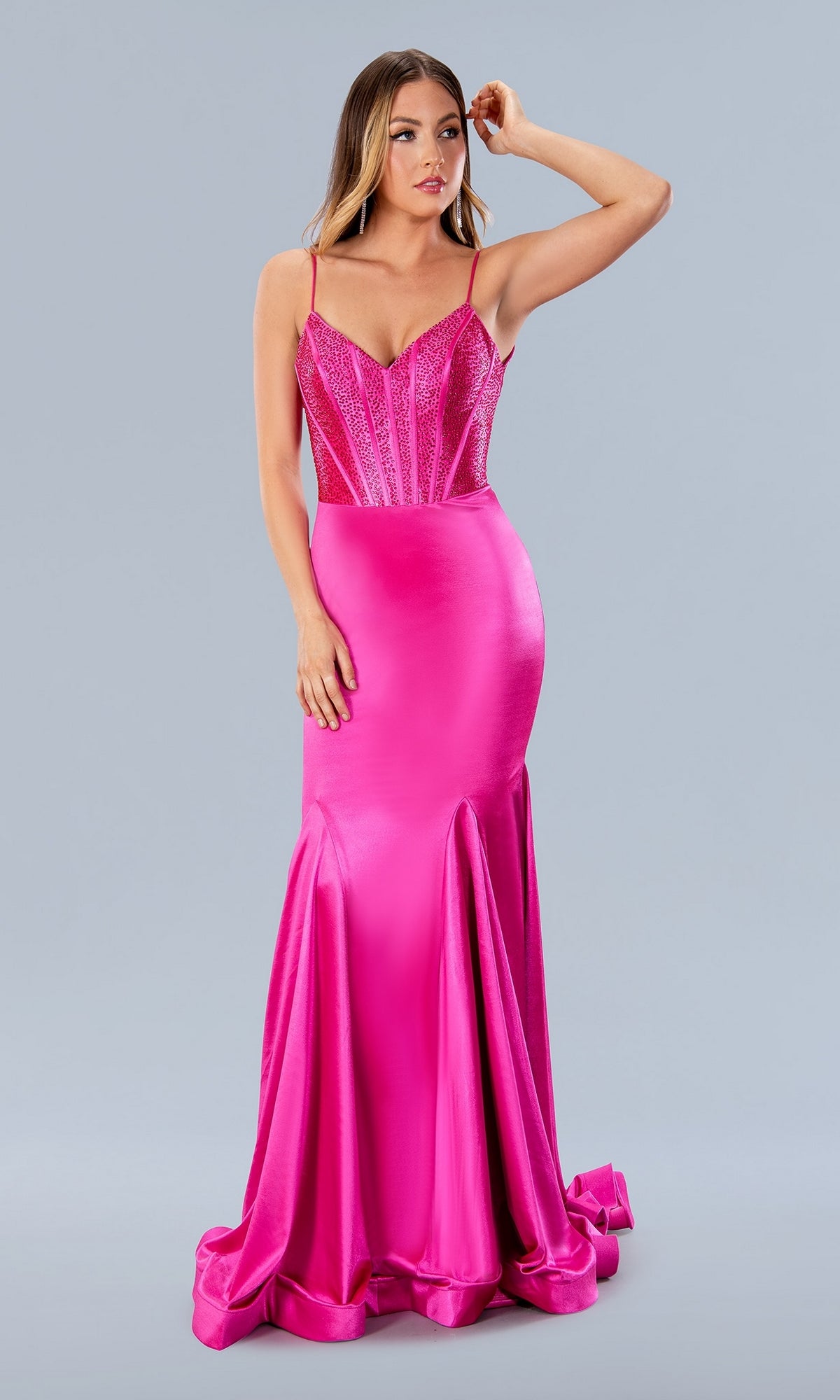 A woman in the Stella Couture 24198 Deep V-Back Long Prom Dress, featuring a bright pink hue, fitted beaded corset bodice, thin straps, and flared ruffled hem, stands against a plain gray background.